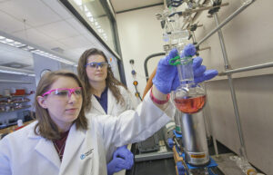 A student holds a beaker filled with a red liquid as a professor looks on.