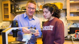 Aleeza Ajmal '18 and James Ferri stand together looking at an object by a microscope in a laboratory.