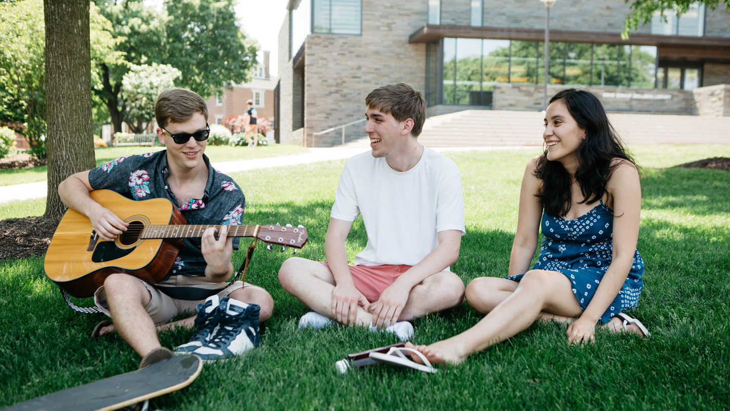 Chris Ulyett '17 plays the guitar on the grass while two friends listen.